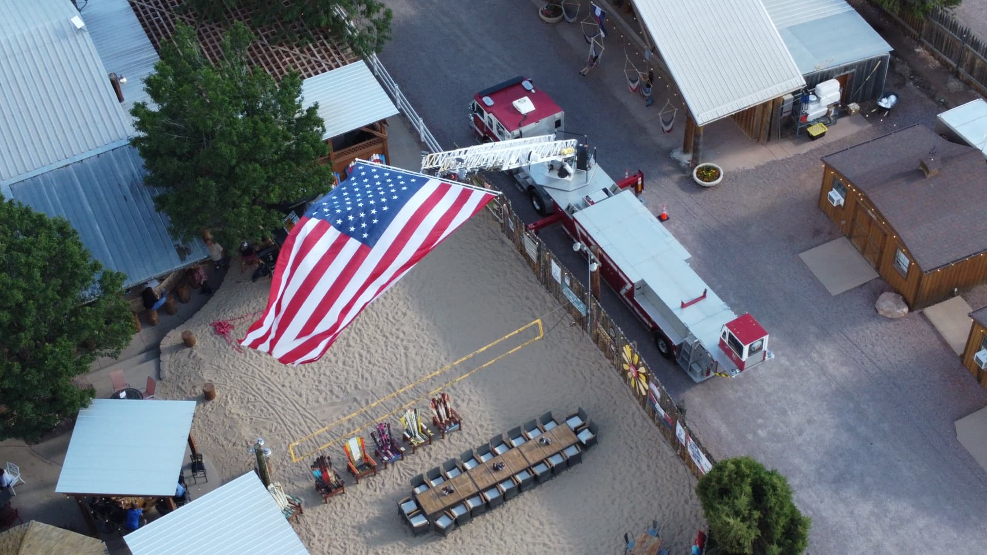 Hero aerial shot — giant American flag suspended from a fire truck ladder over the sand volleyball court, with the full WWBG property, timber pavilion, and Royal Gorge valley in the background.