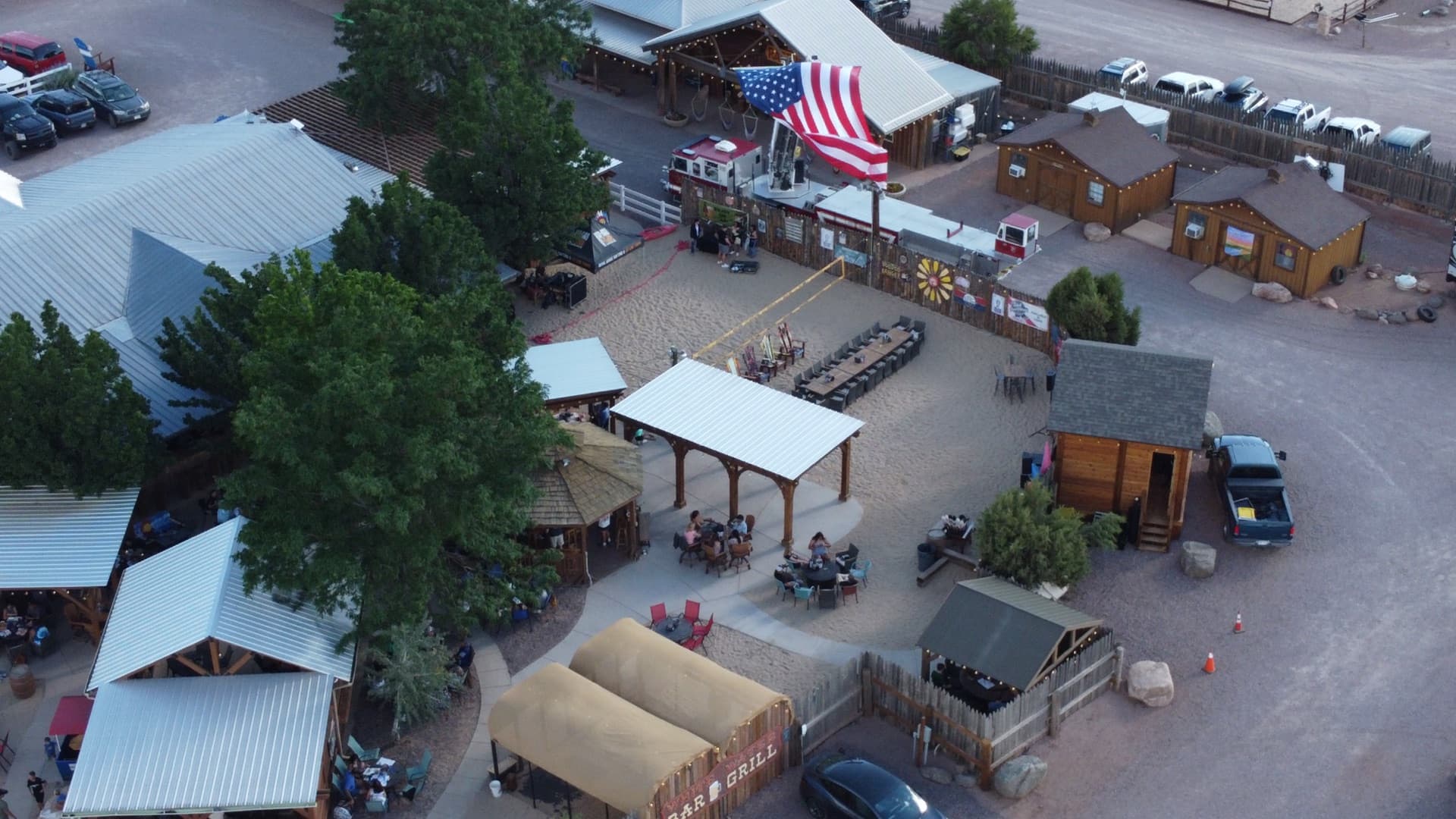 Lower-angle aerial capturing the BAR & GRILL sign, guests dining under the timber pavilion, stage area with live band, string lights, and warm evening atmosphere.
