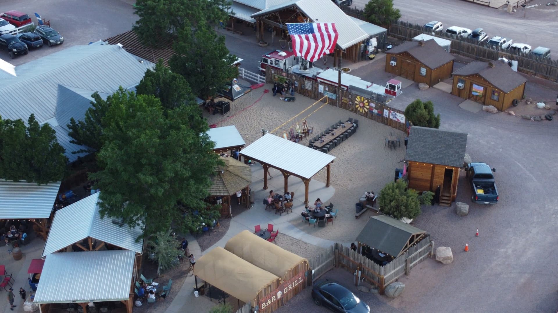 Tighter aerial showing the rustic wooden cabin structures, warm string lights glowing at golden hour, and the American flag overhead.