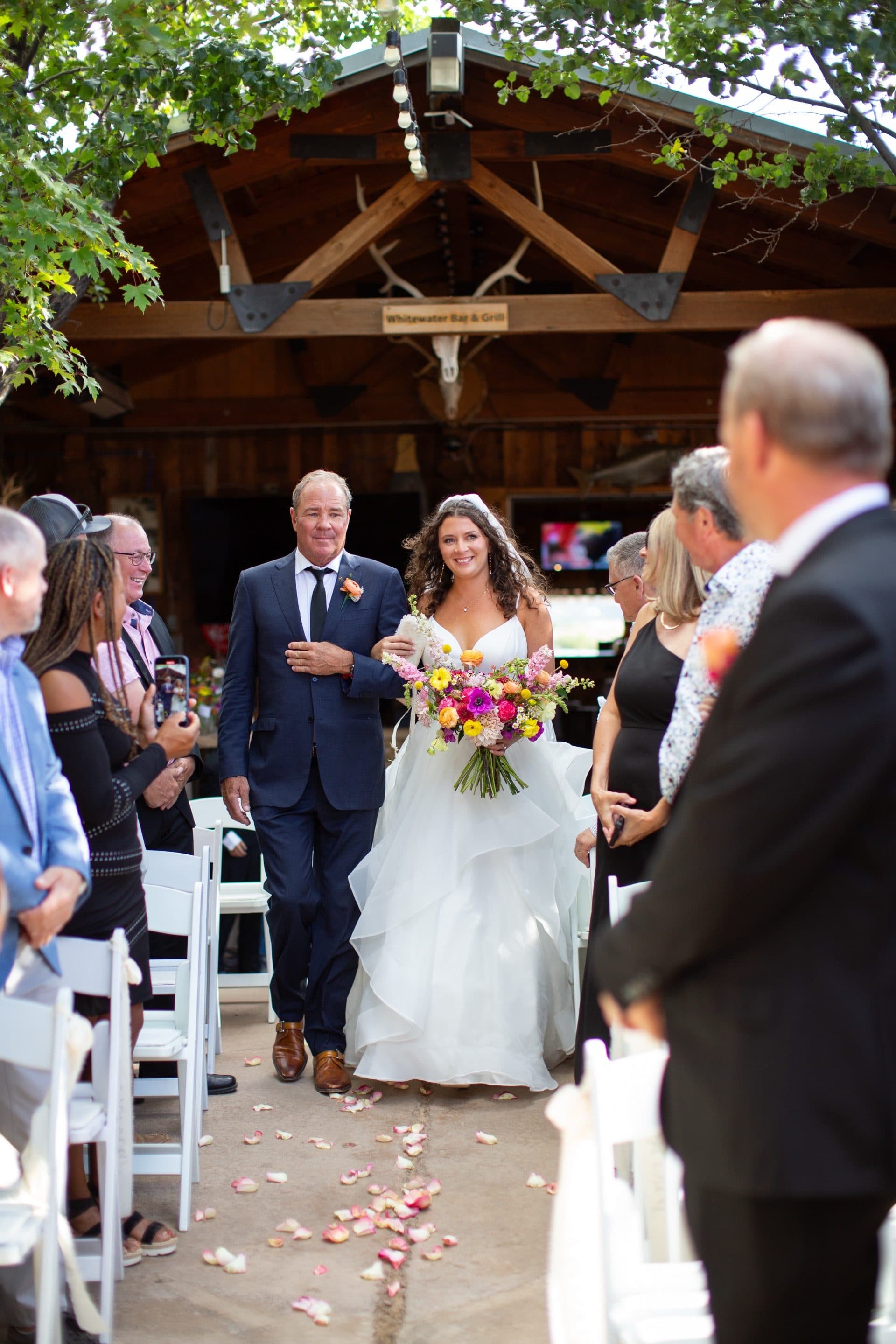 Bride walking down the aisle with her father, carrying a vibrant wildflower bouquet. She wears a flowing ruffled white gown; father in navy suit with cowboy boots. Rose petals scattered on the aisle.