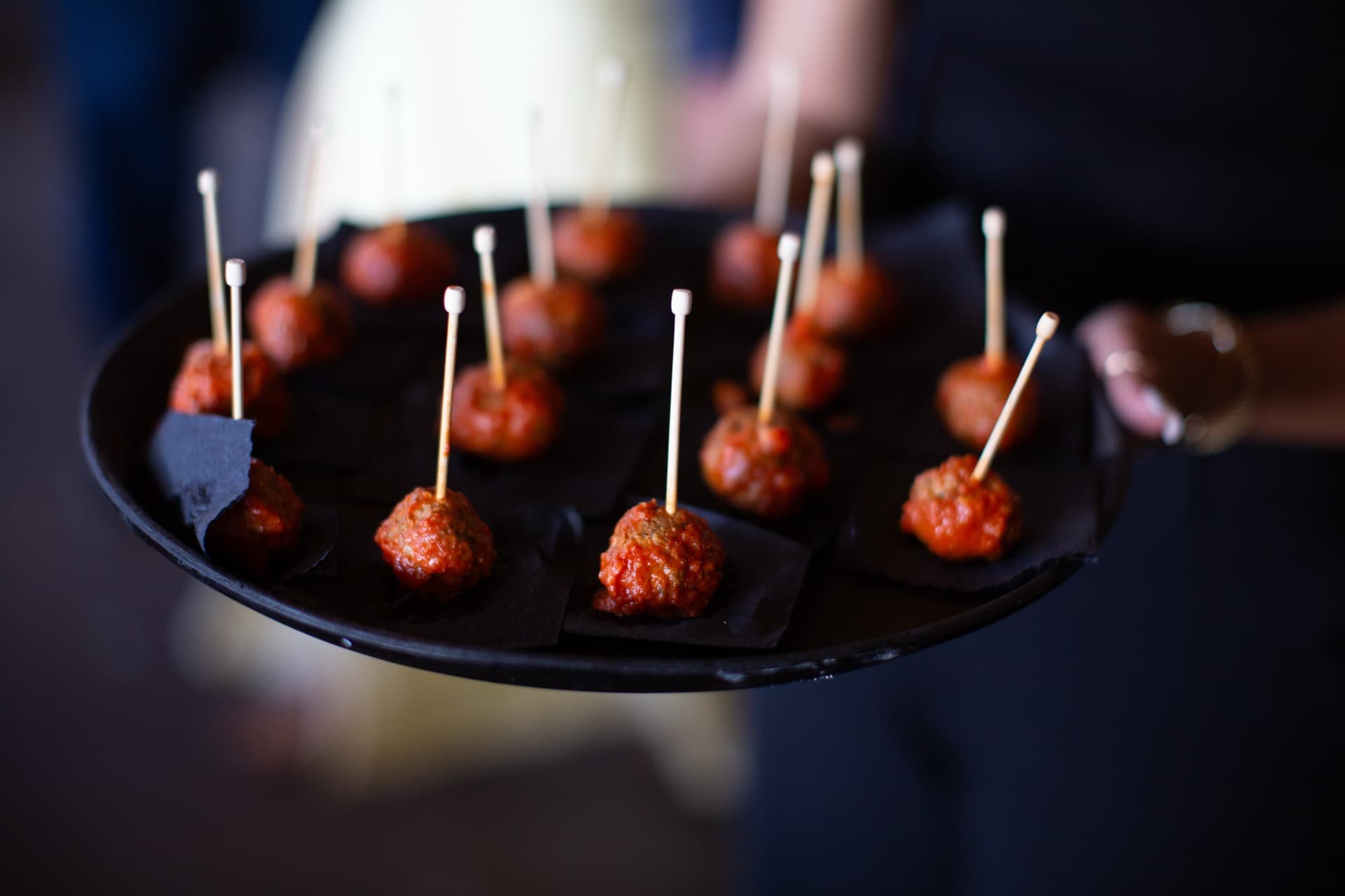 Server holding a black platter of glazed meatballs on toothpicks. Close-up food photography with beautiful bokeh background. WWBG catering quality on display.