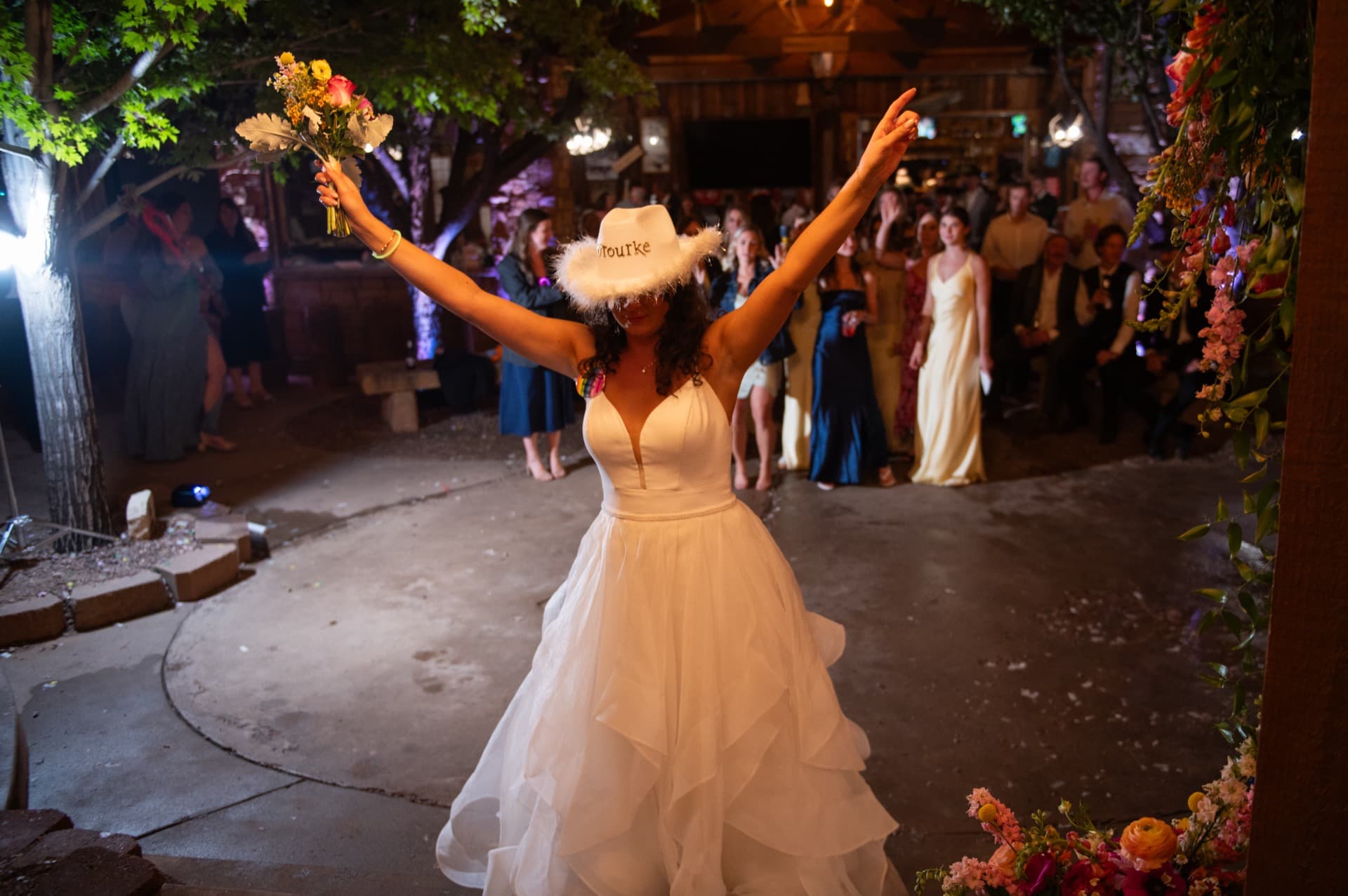 Bride on the gazebo stage about to throw the bouquet. Arms wide holding the colorful wildflower bouquet overhead, wearing a cowboy hat. Female guests gathered below in anticipation.