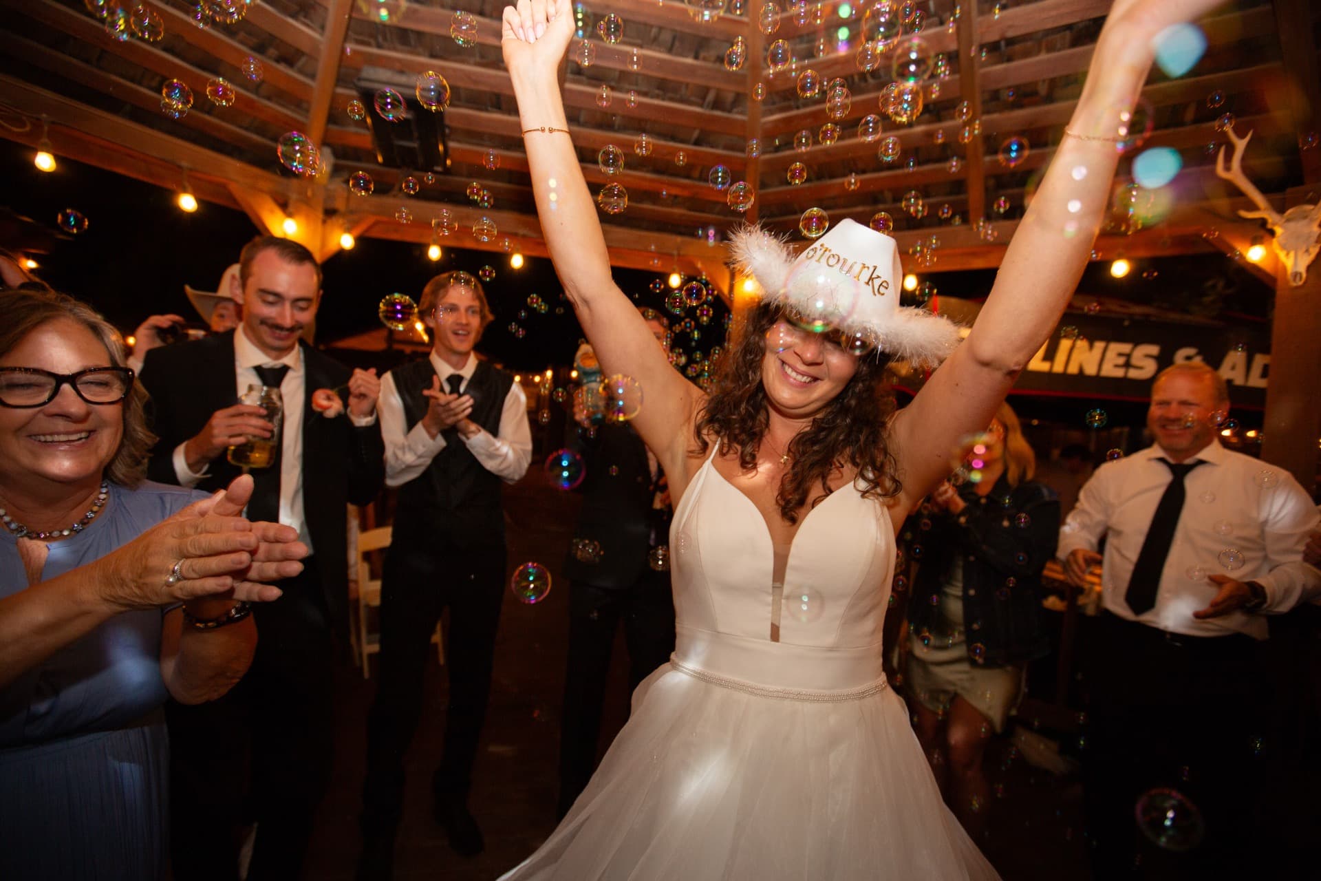 Bride celebrating on the dance floor arms raised, wearing a cowboy hat with feather trim. Bubbles fill the air as guests cheer around her inside the WWBG main space.