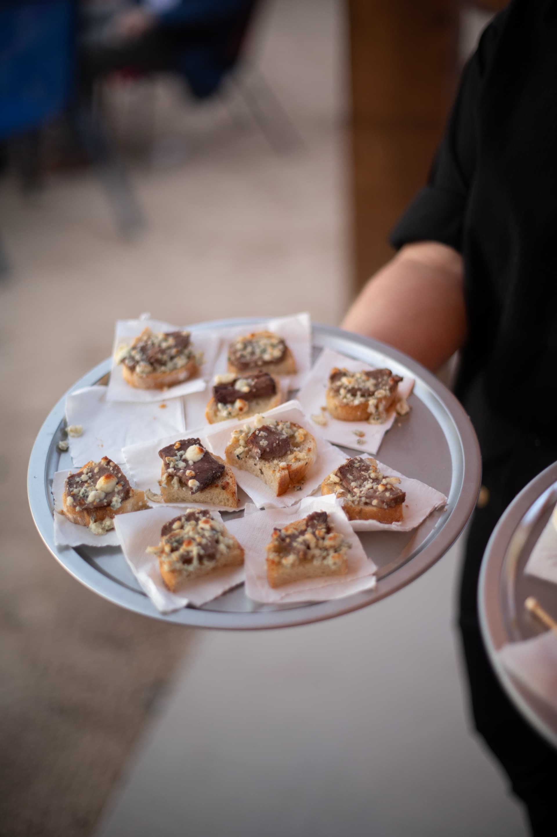 Server holding a silver tray of bruschetta topped with smoked meat and blue cheese crumbles. Artisan quality presentation from WWBG catering.