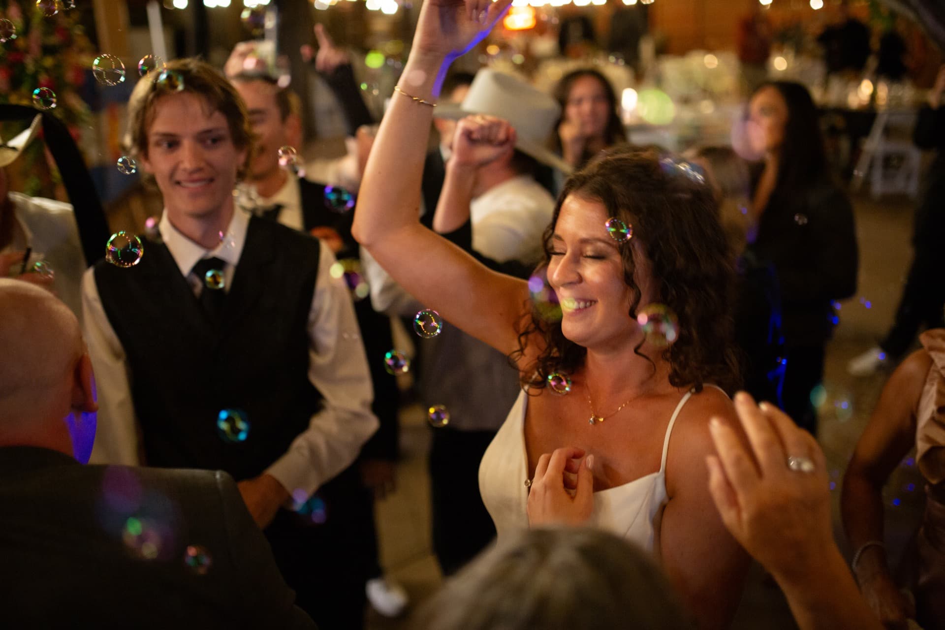 Close-up of bride dancing — bubbles catching rainbow light float around her face. Groom beside her. Beautiful iridescent bubble photography with warm venue lighting.