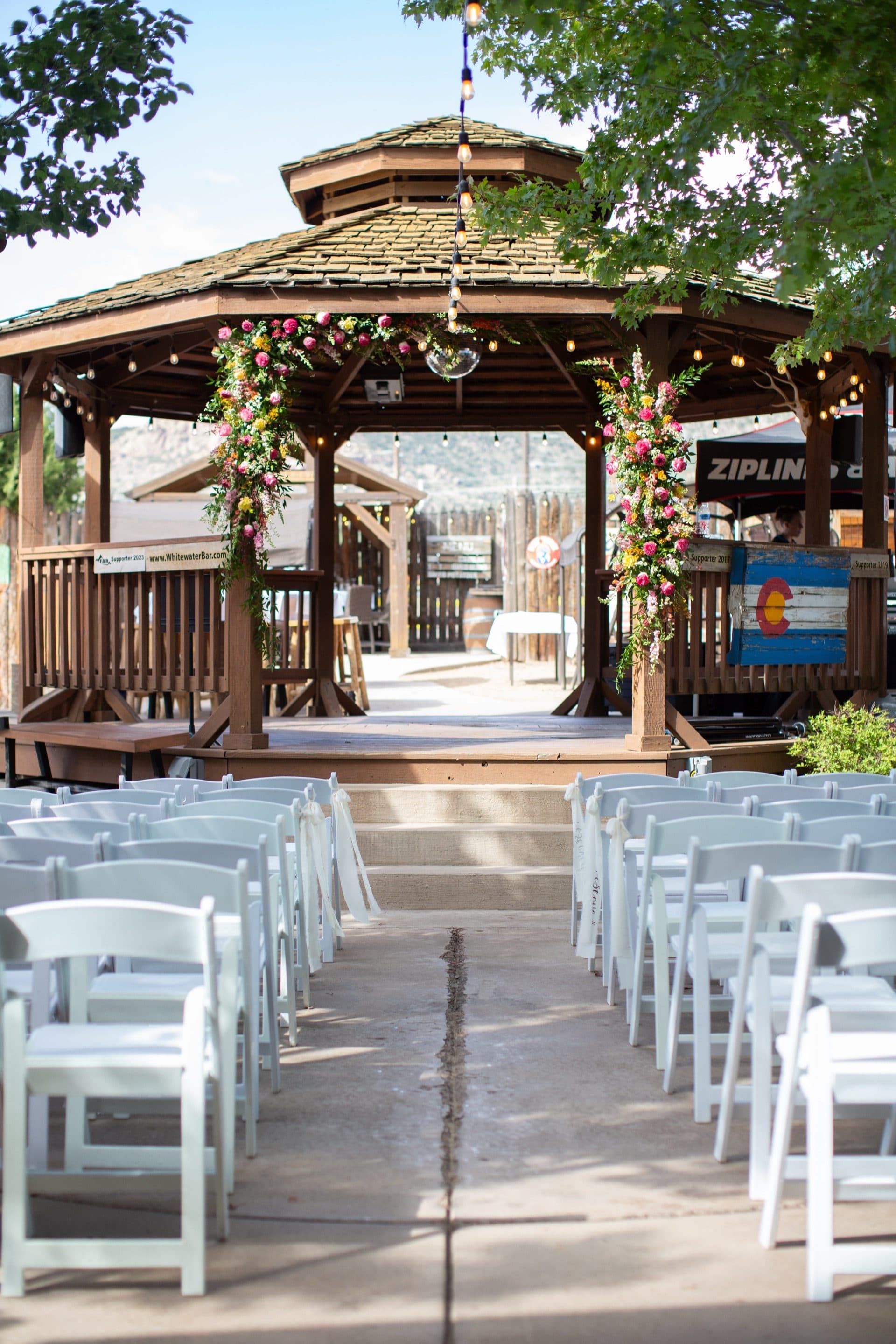 Looking down the aisle toward the WWBG gazebo decorated with a stunning floral arch — pink roses, yellow flowers, and trailing greenery. White folding chairs in neat rows with string lights overhead.
