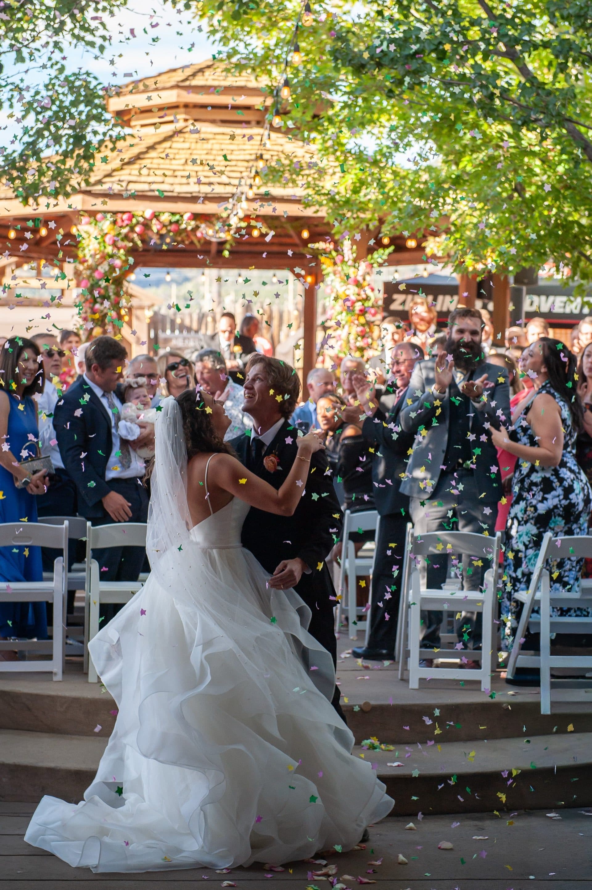 The couple walks back down the aisle as guests shower them with confetti and flower petals. The gazebo with floral arch visible behind — pure joy and celebration.