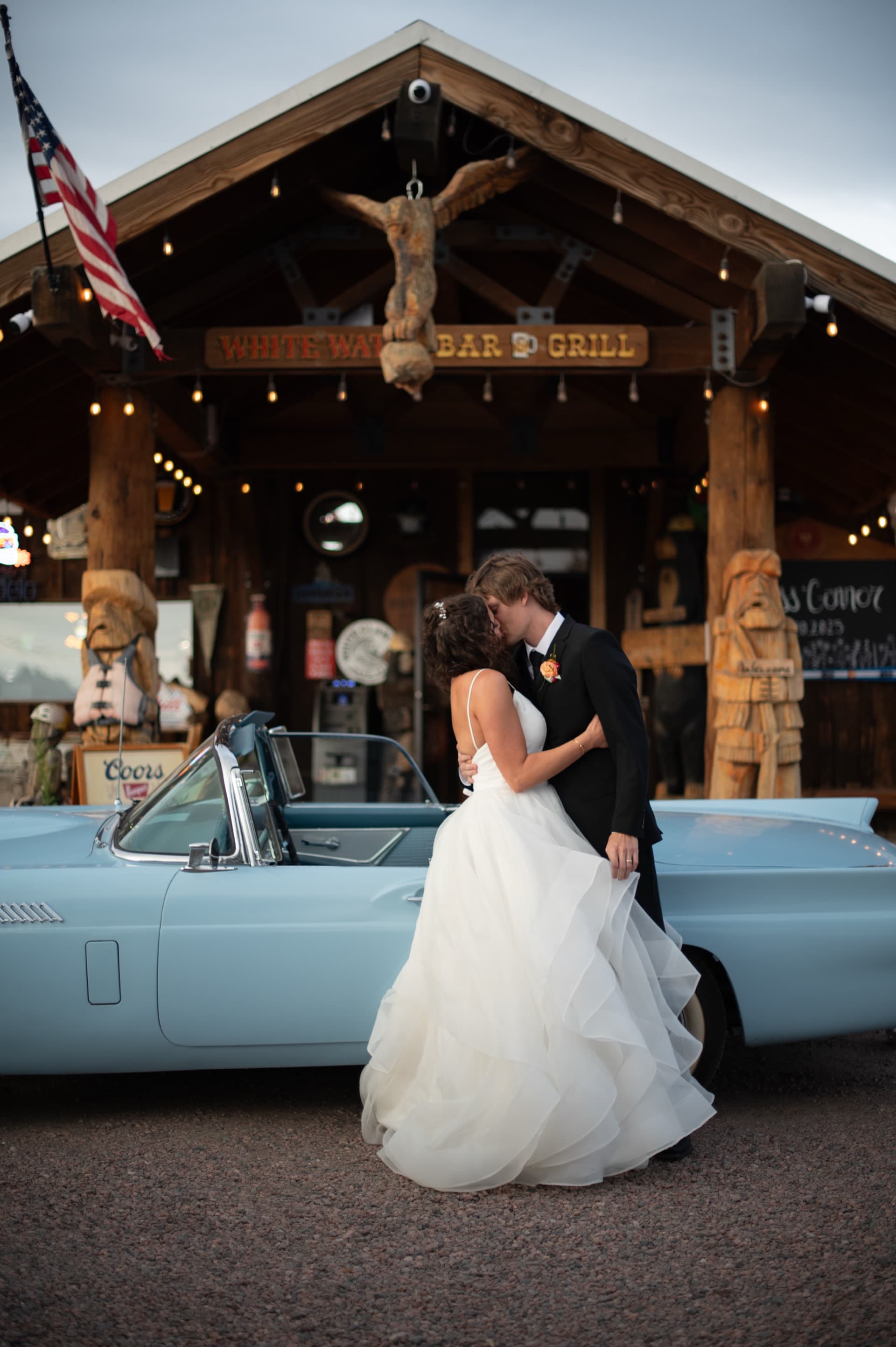 Bride and groom kissing in front of the vintage baby blue 1957 Thunderbird, with the full Whitewater Bar & Grill entrance behind them at dusk. Carved totems, American flag, string lights, and antler mount frame the scene. The definitive WWBG wedding photo.