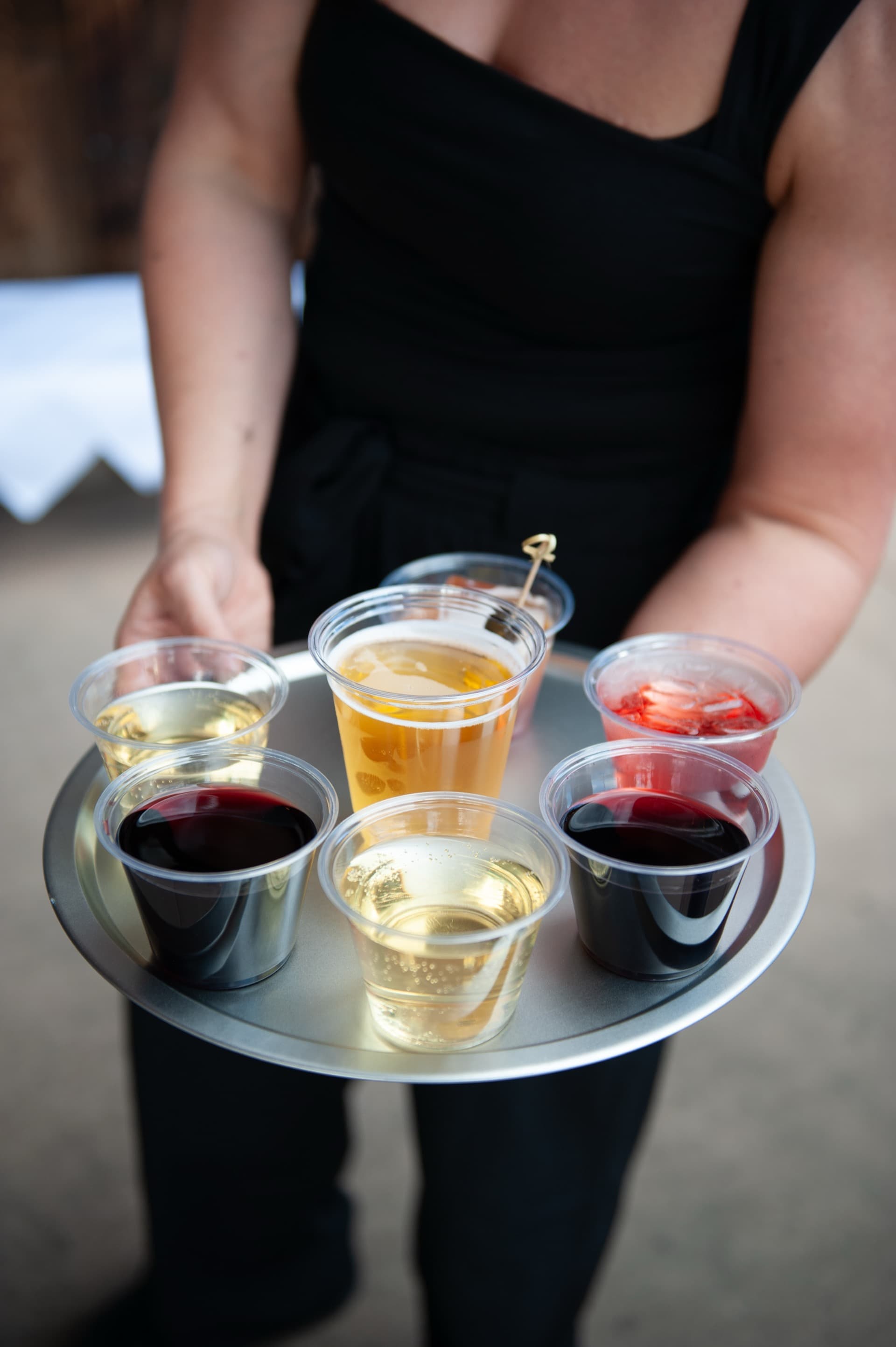 Server holding a round silver tray of drinks — red wines, white wines, champagne, craft beer, and a strawberry cocktail. The full WWBG bar service on display.