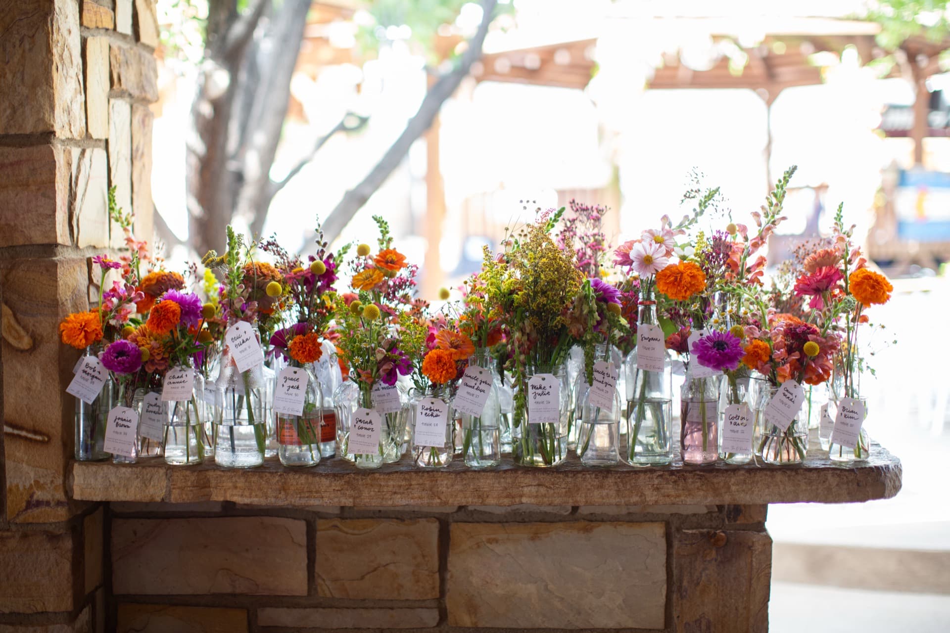 Dozens of glass bottles and mason jars filled with vibrant wildflowers on the WWBG stone fireplace mantel. Each bottle has a handwritten calligraphy tag with guest names and table assignments.