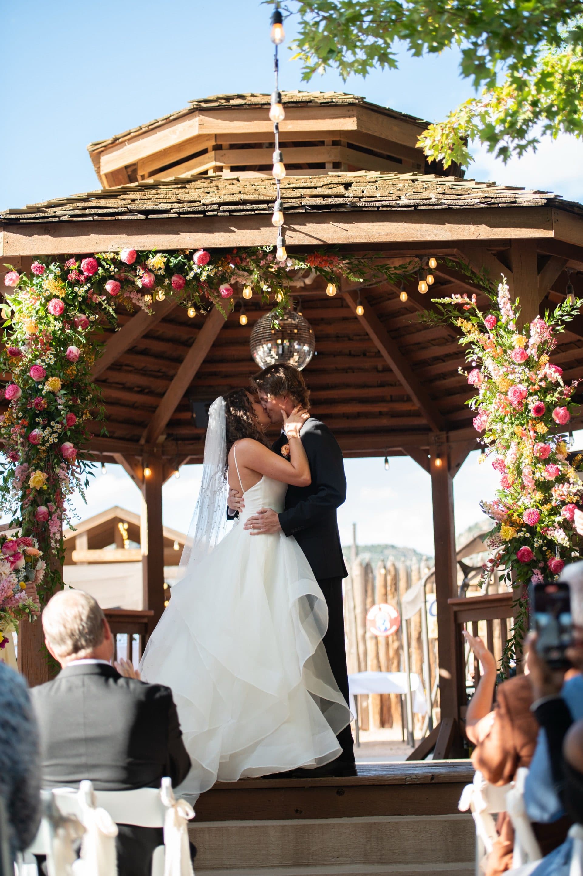 Bride and groom share their first kiss on the gazebo stage under the floral arch. String lights and blue Colorado sky behind. Guests' phones capture the moment in the foreground.