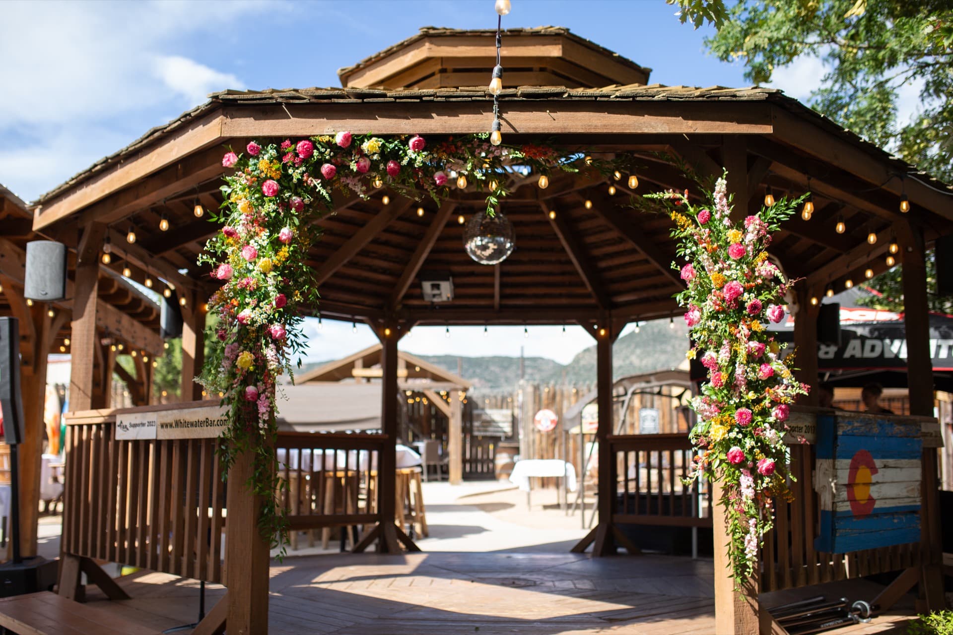 Close-up of the gazebo floral arch from below — roses, ranunculus, dahlias, and greenery cascading over the timber arch. String globe lights overhead against blue sky.
