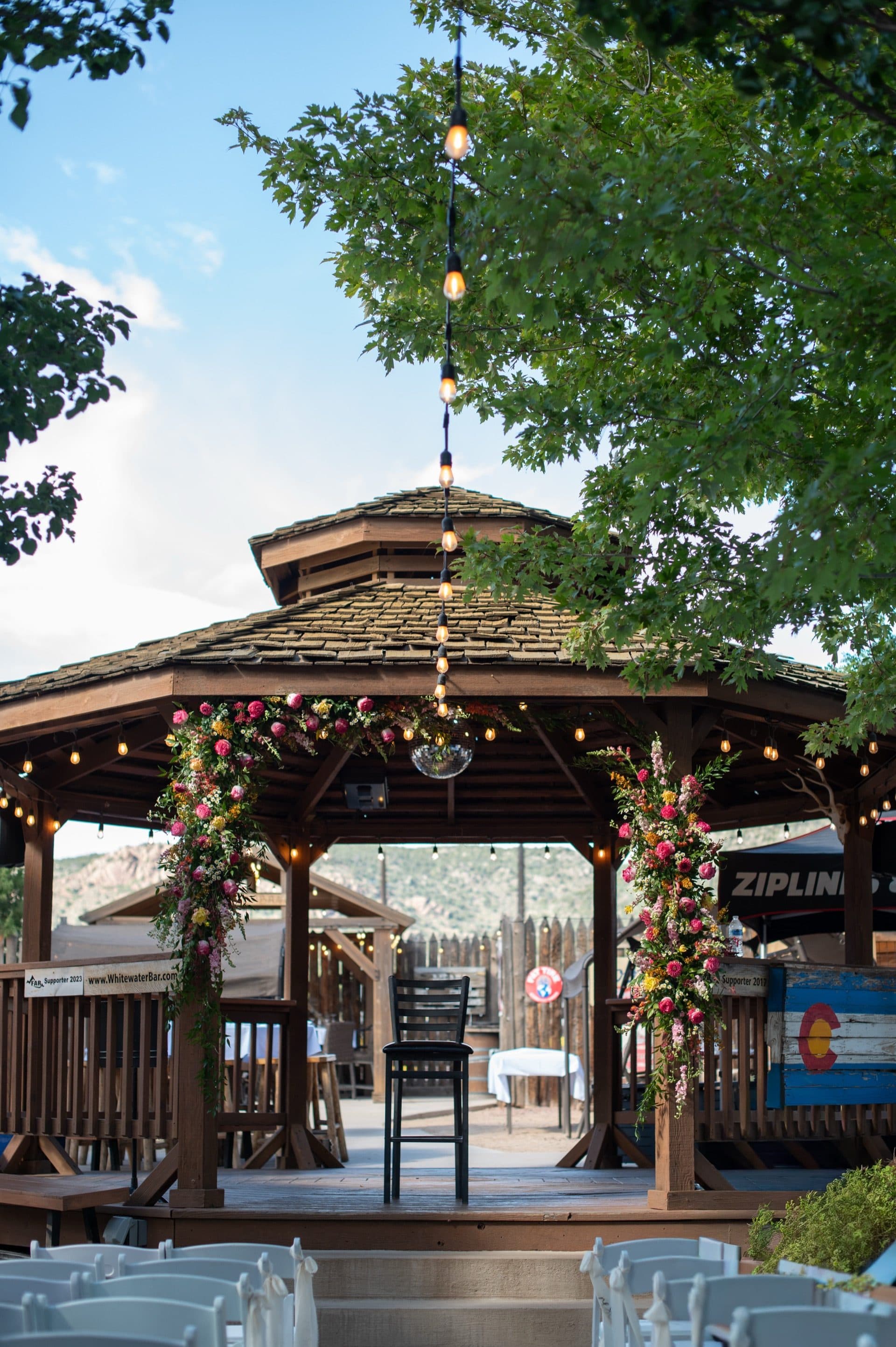 The gazebo at golden hour — floral arch and string lights glow warmly against the evening sky. Timber construction and shingled cupola beautifully lit. Magical atmosphere.