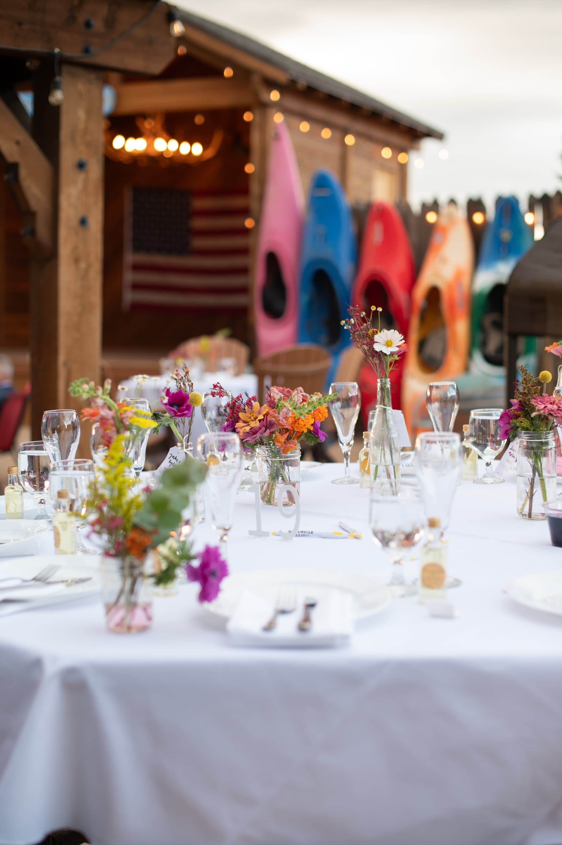 Reception table set with white linens and wildflower centerpieces. Background: colorful kayaks hanging on the WWBG adventure rack with American flag and string lights. The quintessential WWBG wedding shot.