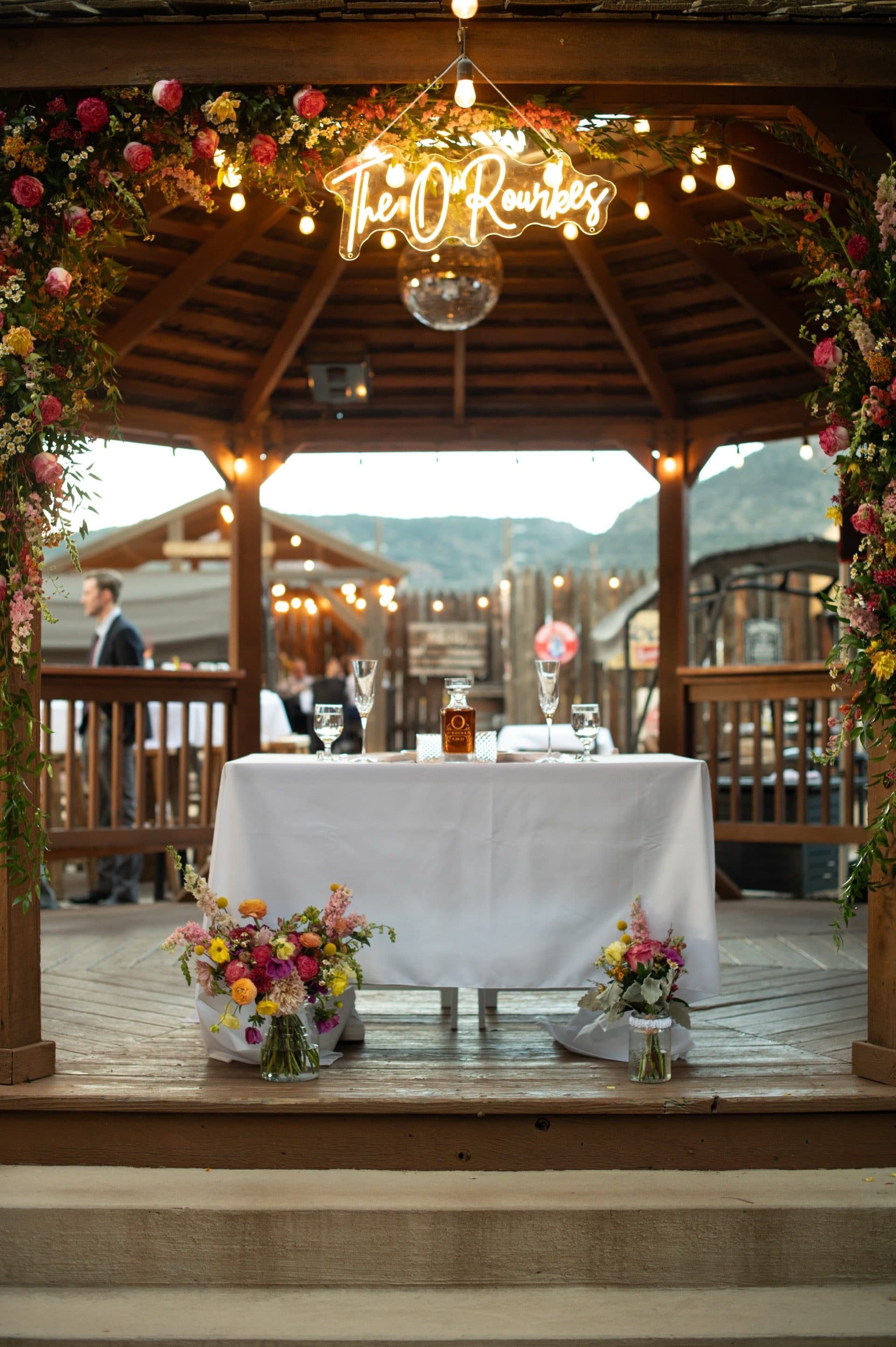 The sweetheart table in the gazebo at dusk with a custom neon sign glowing above. Floral arch frames the intimate setting with string lights overhead and mountains visible through the gazebo.
