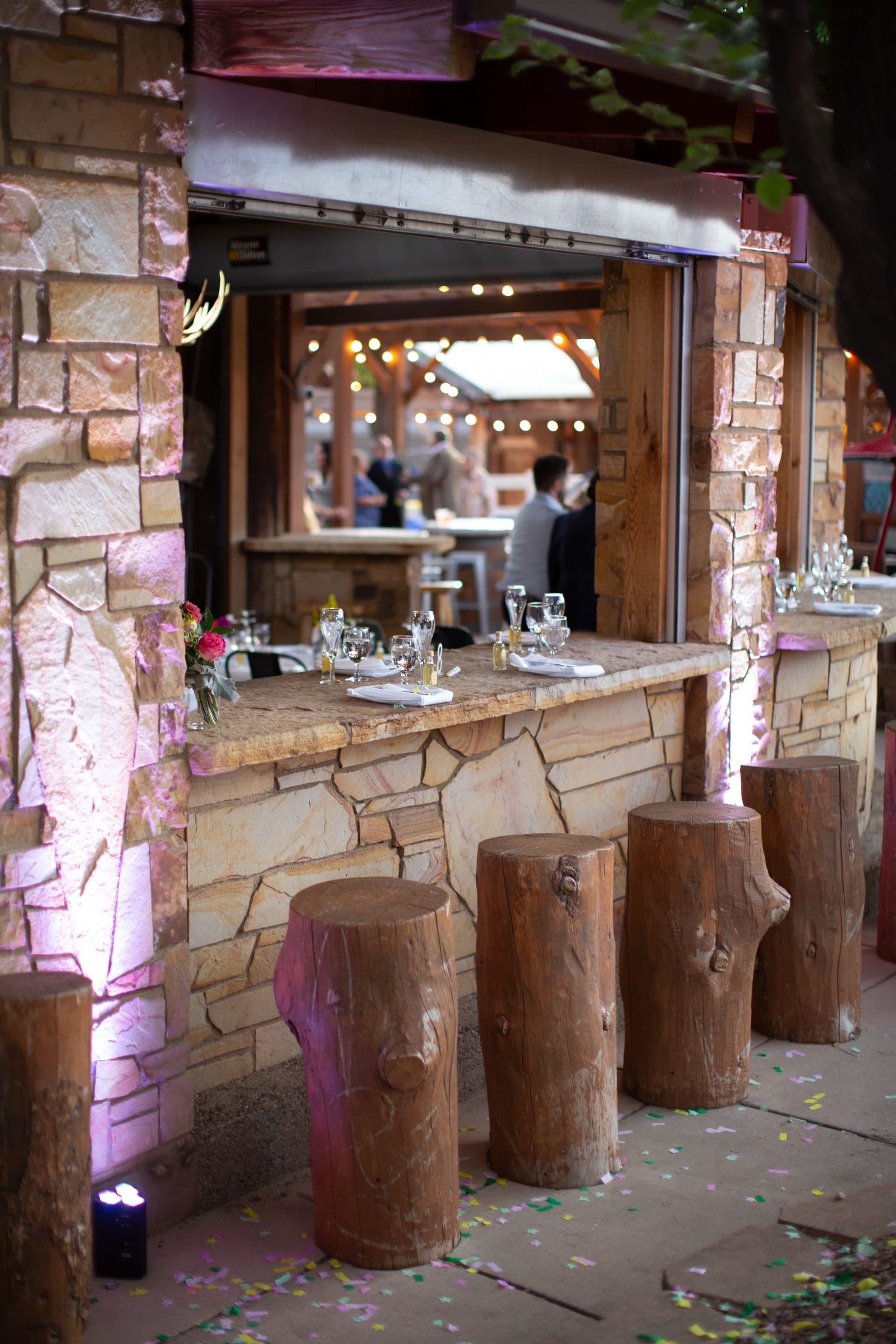 WWBG outdoor stone bar at night with purple uplighting. Log-stump bar stools line the stone counter. Confetti scattered on the ground. Evening ambiance at its finest.