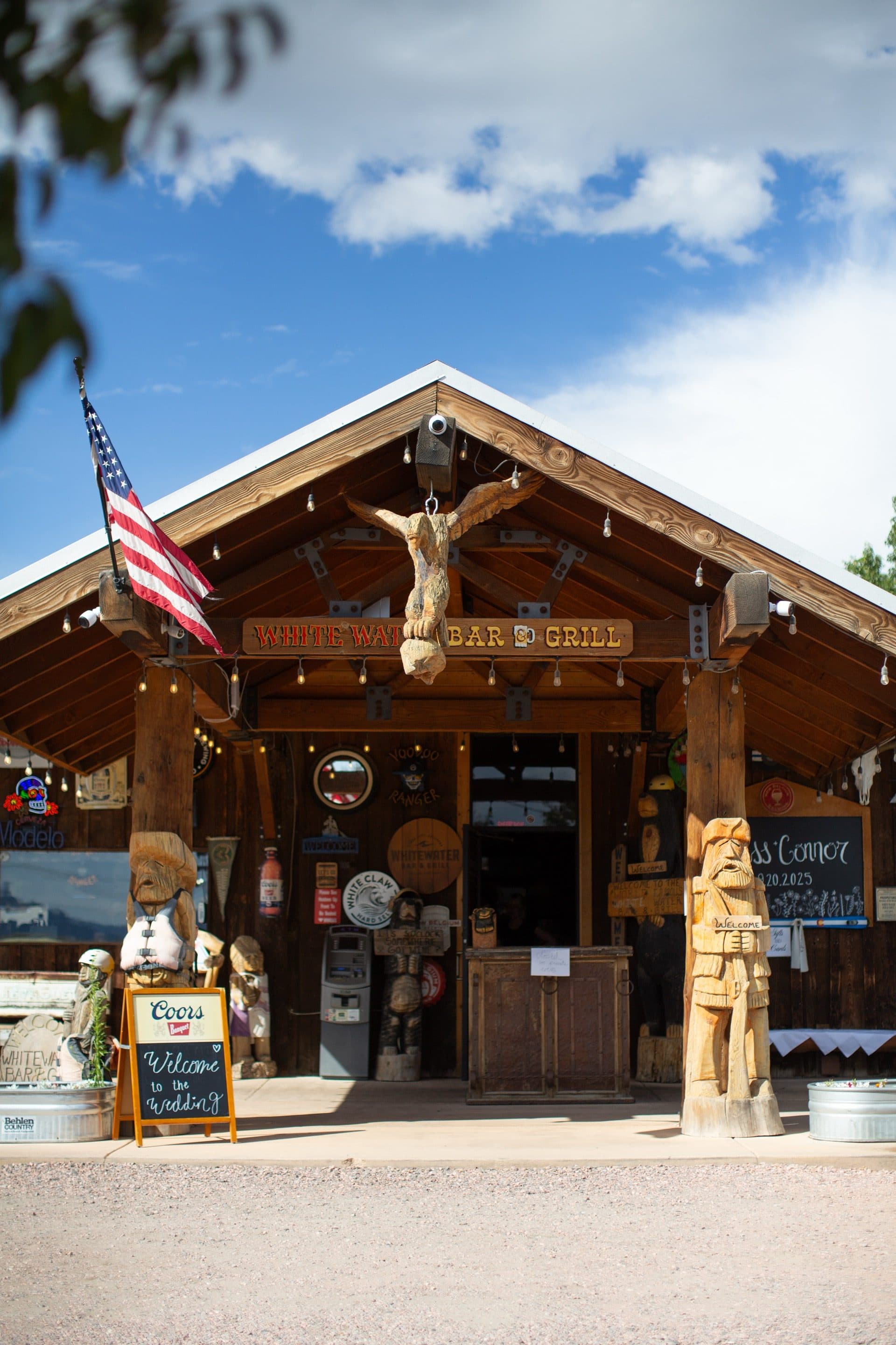 WWBG front entrance exterior in daylight. Rustic log cabin architecture with peaked roof, carved wooden totem figures, antler mount above the sign, American flag, and string lights.