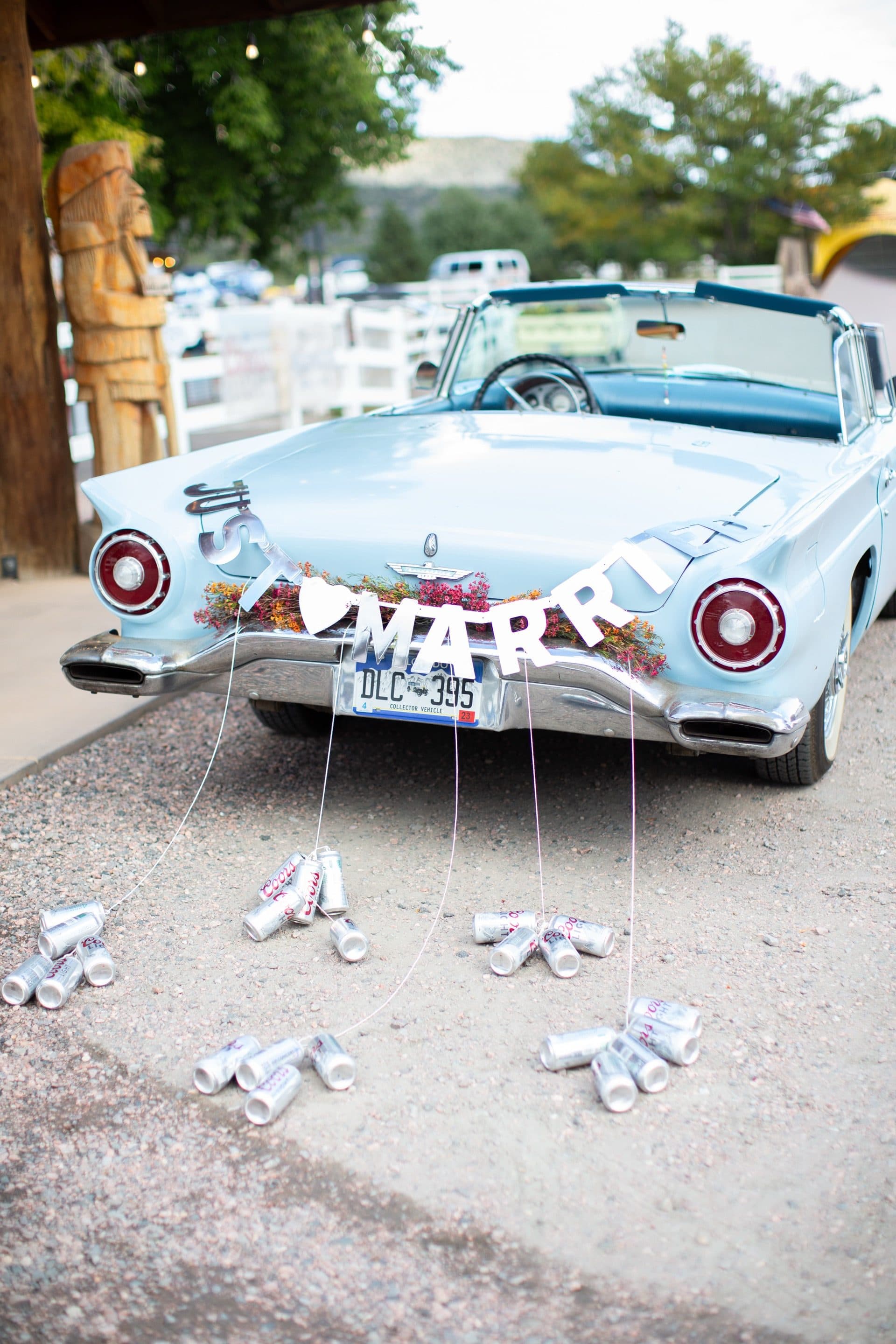 Vintage baby blue 1957 Ford Thunderbird convertible with "MARRIED" letter banner and tin cans tied to the bumper. Carved wooden totems visible in background. Classic Americana wedding exit vehicle.